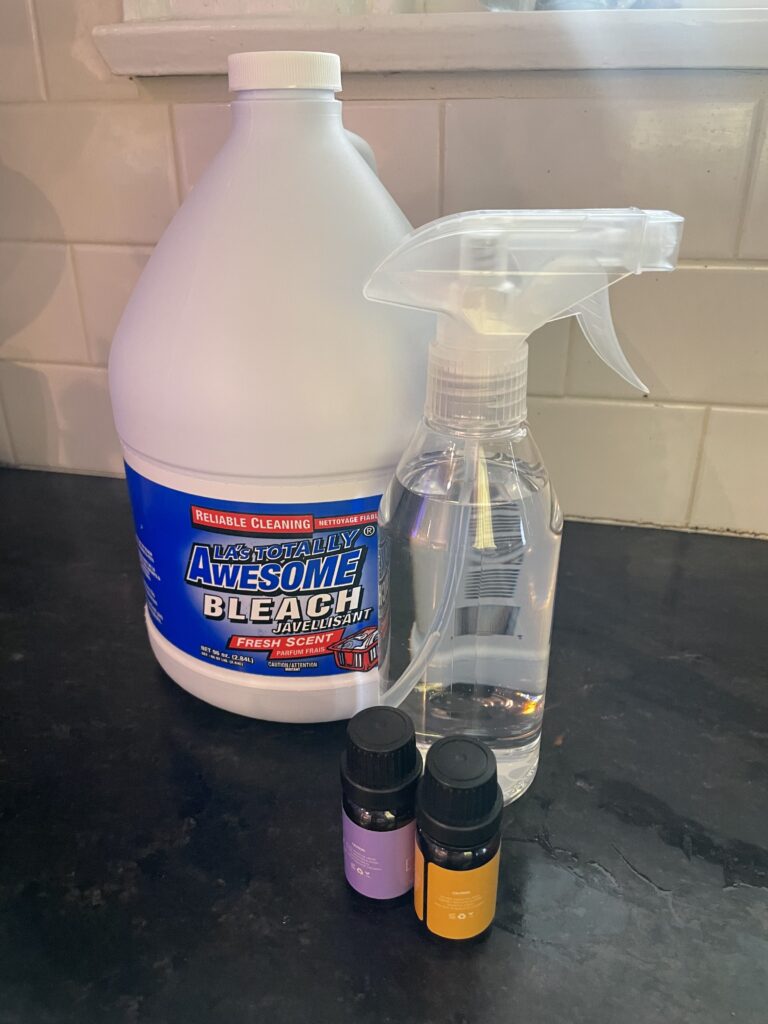A spray bottle filled with diy disinfectant, a bottle of bleach, and essential oils on a countertop. 